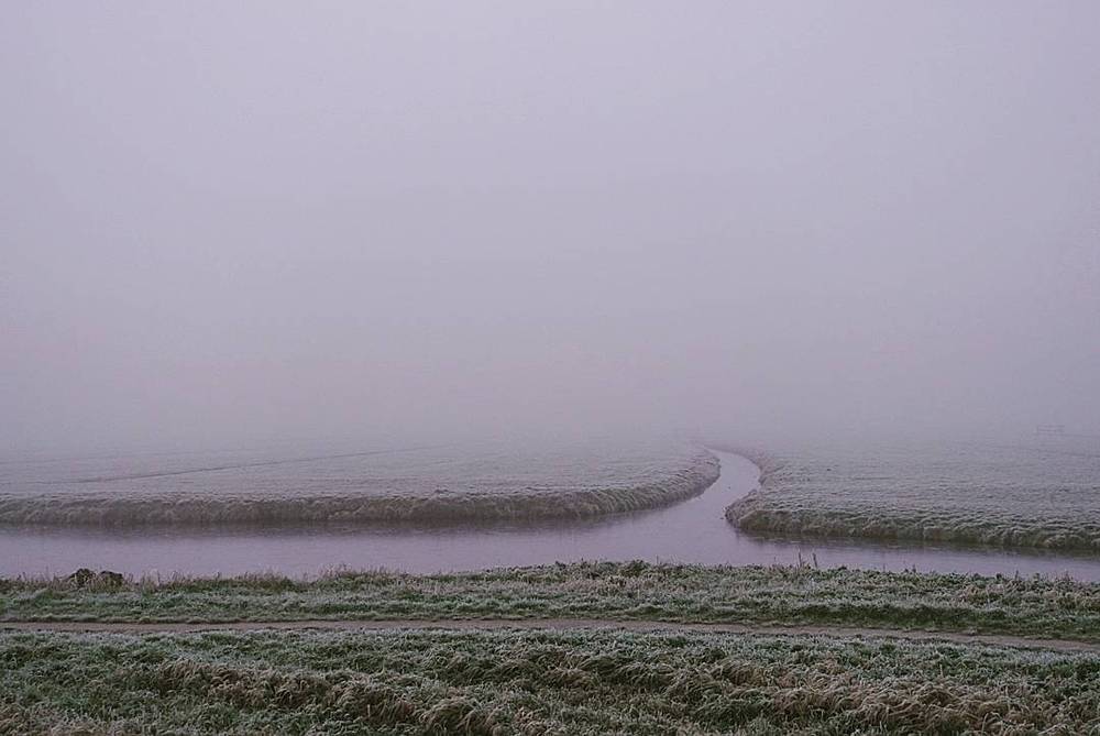 A winding river through a frost-covered field shrouded in fog.