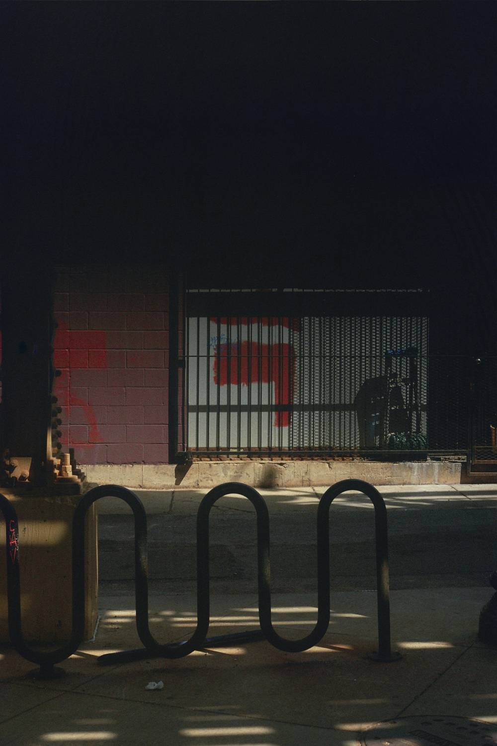 Bike rack in the foreground under an elevated train station, a wall in the background with splotches of red paint. Similar to the first image, the top half of the image fades to black due to a film issue