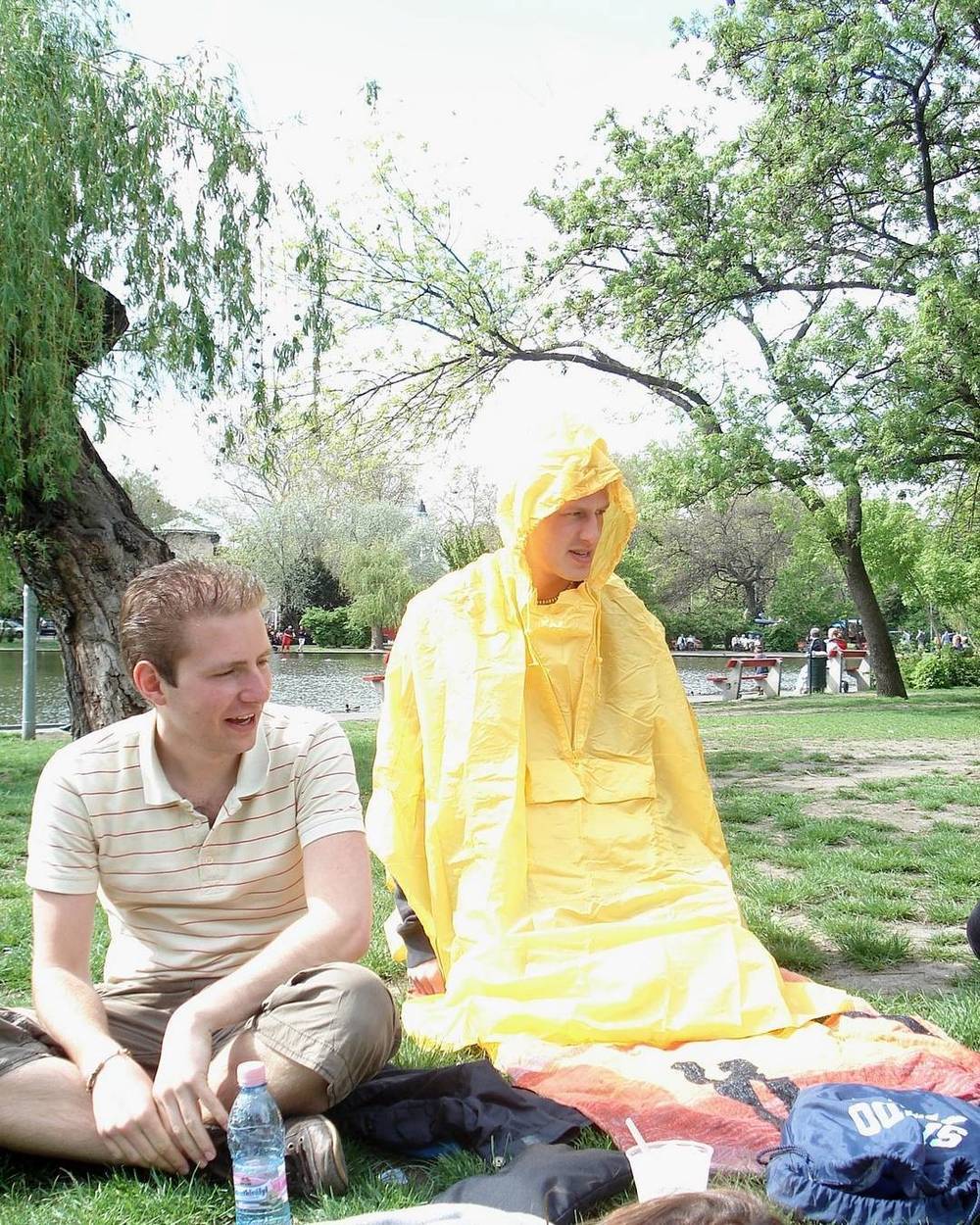 An old photo of two people sitting on grass in a park, one wrapped in a yellow rain poncho.