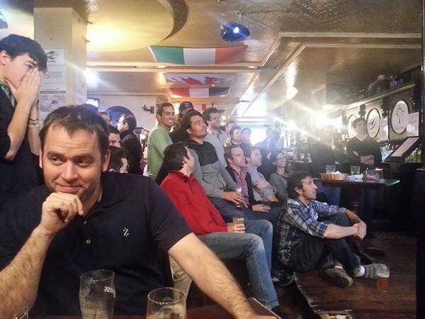 A crowd gathers inside a bar decorated with flags, watching something off-camera.