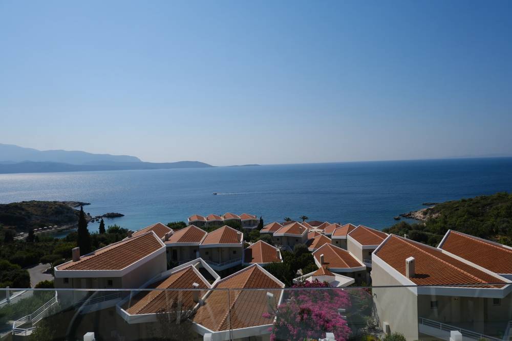 top-floor (restaurant!) view over a coastal resort with orange-tiled rooftops and pink-flowering bushes, overlooking a bay with Turkey hazy in the distance
