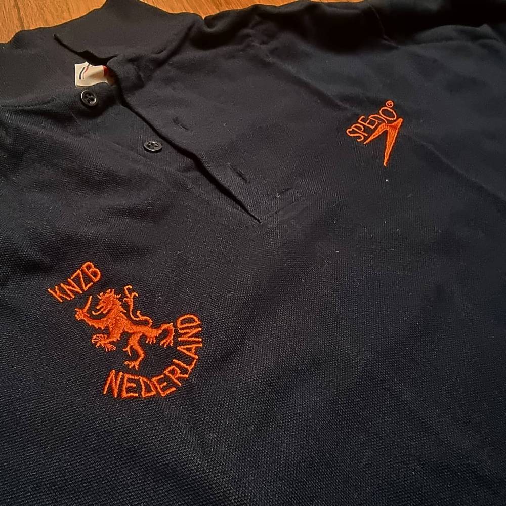 A black polo shirt embroidered with the orange KNZB Nederland swimming federation lion logo.