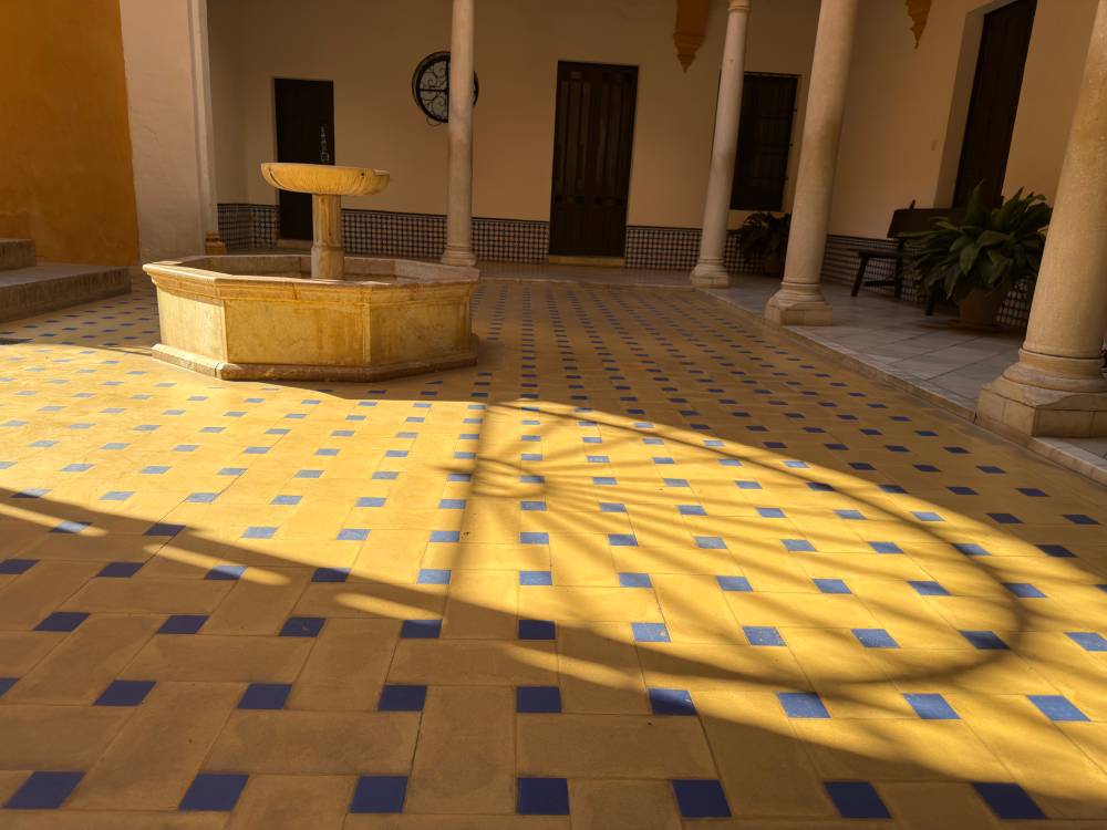 First floor of an open courtyard with a fountain in the middle. The floor tiles are yellow and blue. There is a shadow on the floor of a rounded geometric pattern.