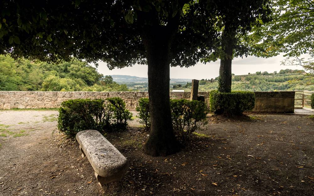 A weathered stone bench beneath a shade tree on a gravel terrace, overlooking a low stone wall with rolling countryside stretching to distant hills.