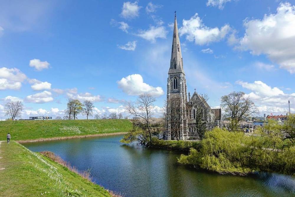 A tall church spire beside a river and green embankment under a blue sky.