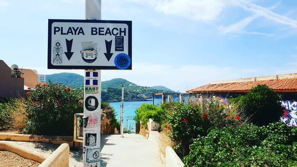A signpost pointing to Playa Beach, surrounded by flowering shrubs and sea views.